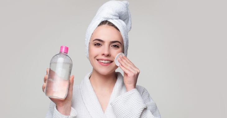 Young woman wearing a bathrobe and a towel over her hair applying miceral water on face using a cotton pad. 
