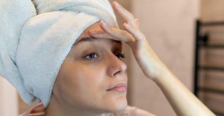 A concerned woman with a towel over her hair looking at the skin on her eyelids in the bathroom mirror